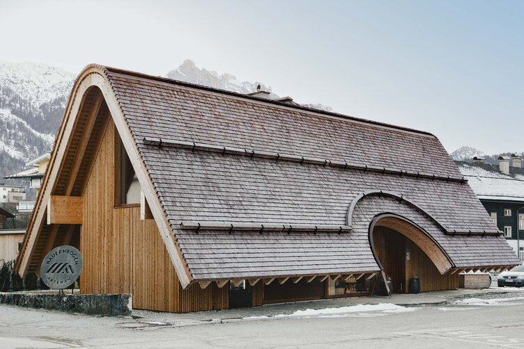 Bauernbogen Abtenau – Außenansicht mit geschwungenem Bogendach und vertikaler Holzfassade, realisiert mit LIGNOLOC® Holznägeln von BECK